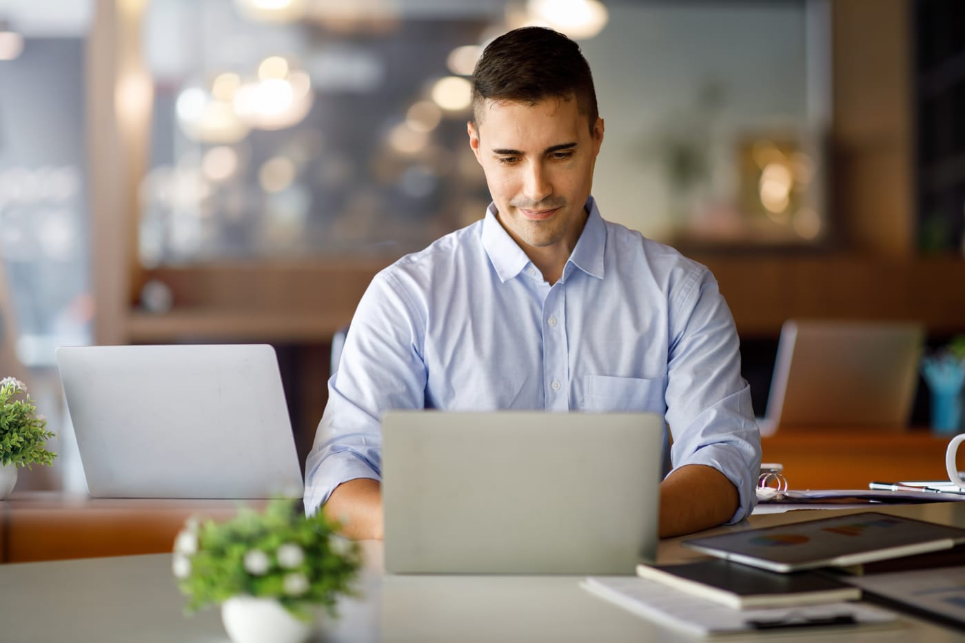 A man working at a laptop in a bright open office, a second laptop and a small plant on the desk beside him.