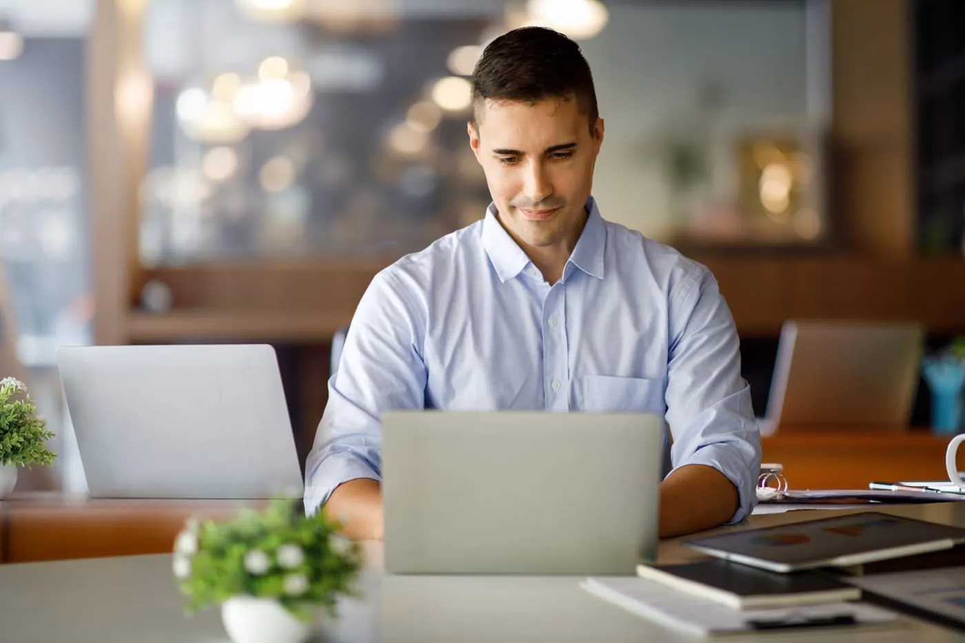 Un homme travaille sur un portable dans un bureau à aire ouverte lumineux, un second portable et une petite plante posés sur la table à côté de lui.