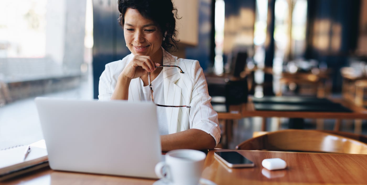 A woman at a laptop in a café, her glasses in hand, a coffee cup and a phone on the table beside her.