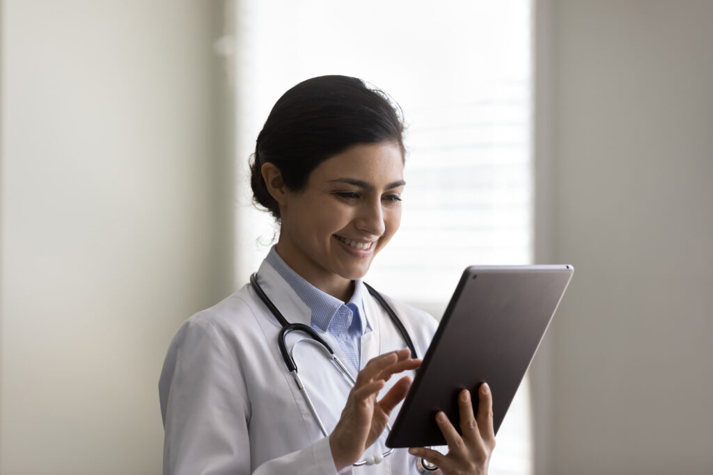 A clinician in a white coat, stethoscope around her neck, reading from a tablet.