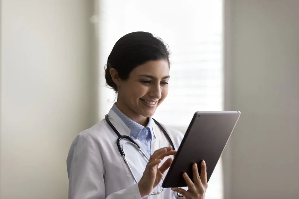 Une clinicienne en sarrau, stéthoscope au cou, qui consulte une tablette.
