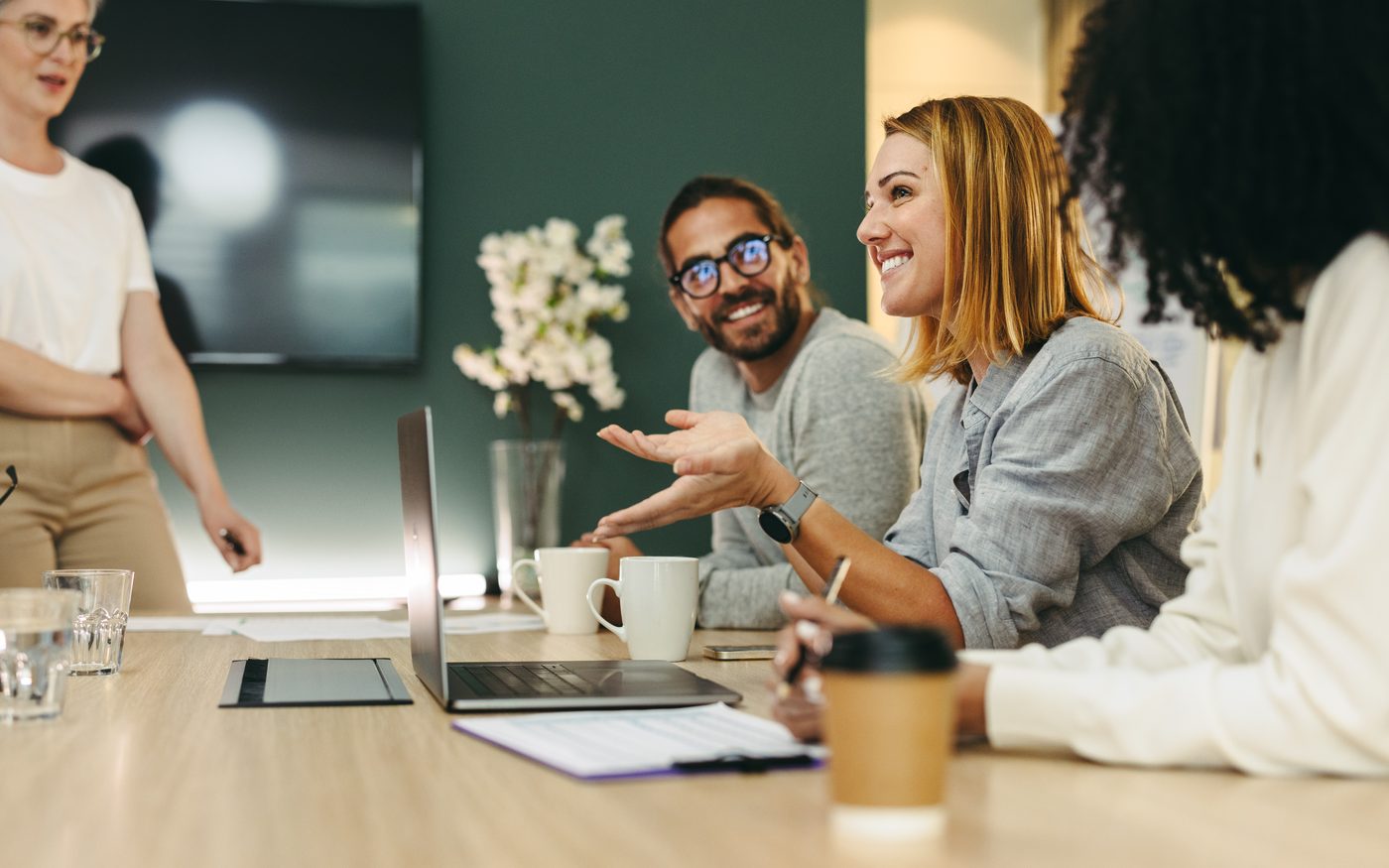 Colleagues around a meeting table, mid-discussion in front of an open laptop.