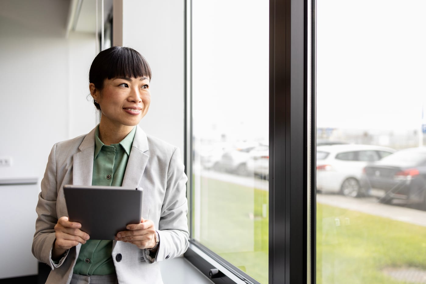 A manager standing at an office window with a tablet, looking out.